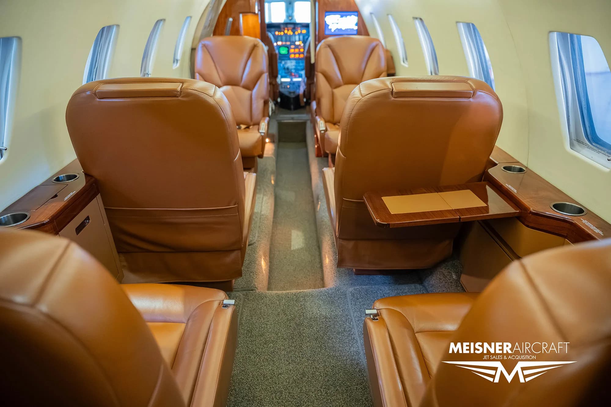 The interior cabin of a private jet with brown leather seats and small side tables, viewed from the rear.