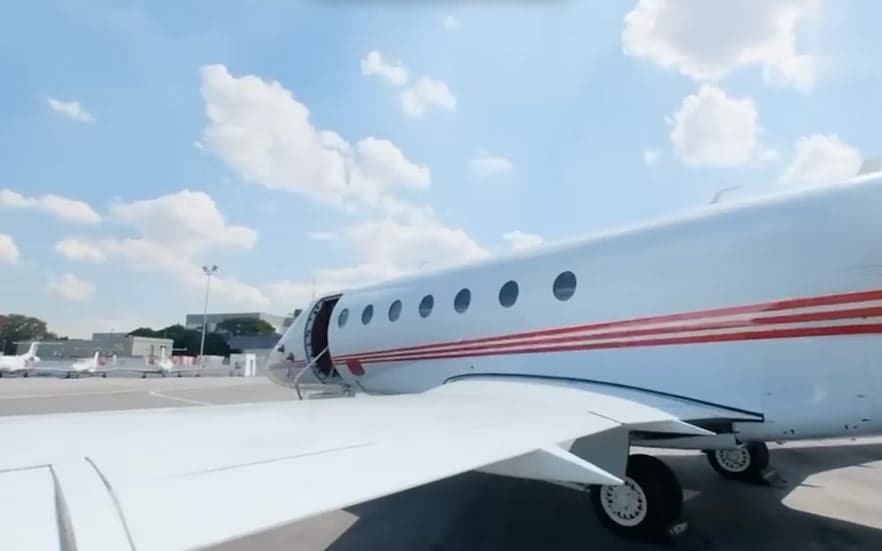 A side view of a white business jet with red stripes, showing the wing and fuselage with the main door open, parked on an tarmac.