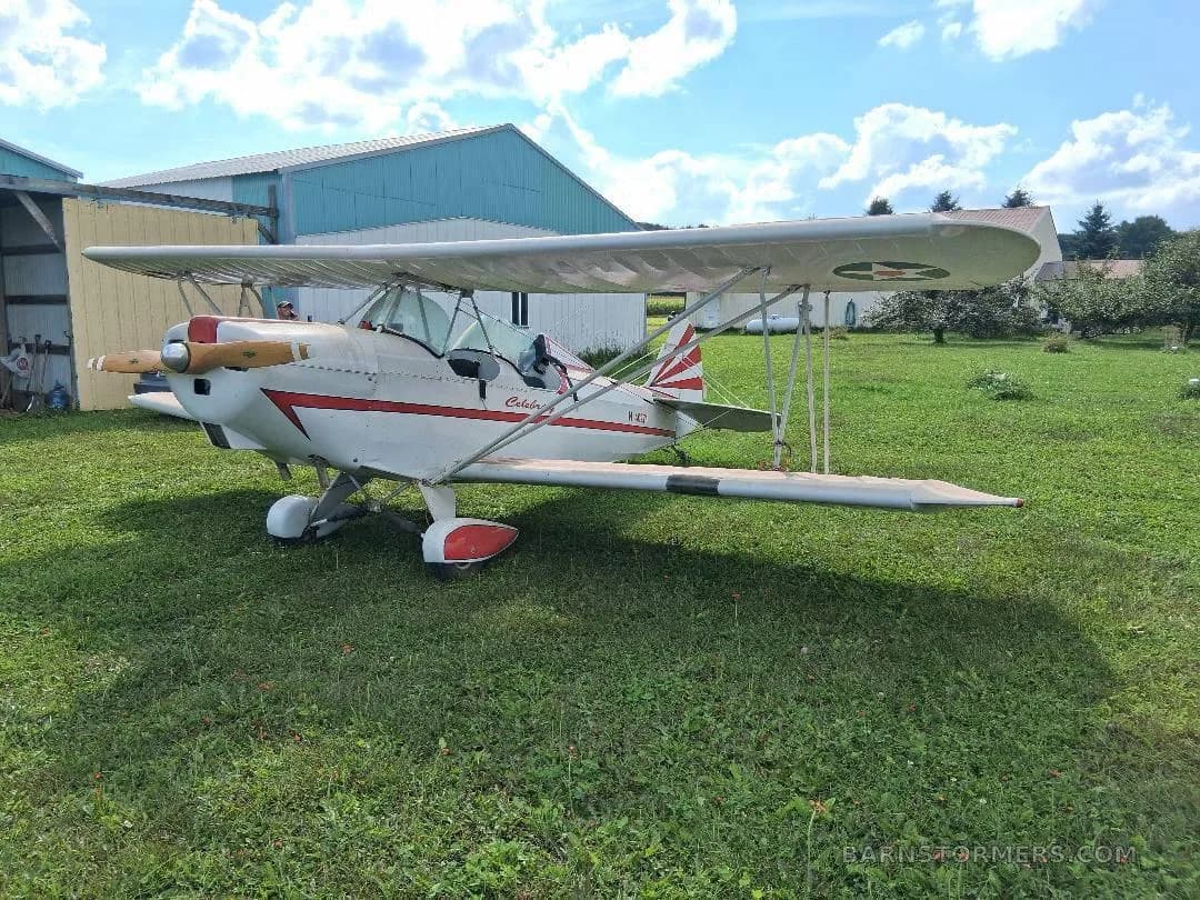 A white biplane with red stripes and a wooden propeller, viewed from the left side, parked on green grass next to a blue hangar.