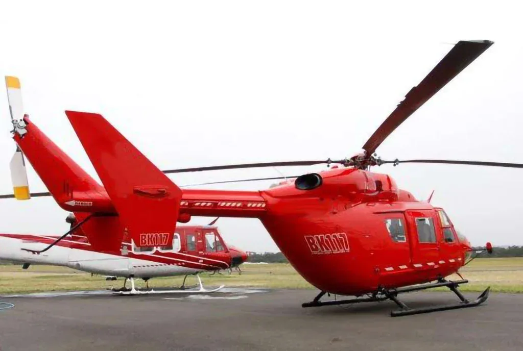 A red BK117 helicopter with 'BK117' on its side, parked on an airfield with another helicopter behind it.
