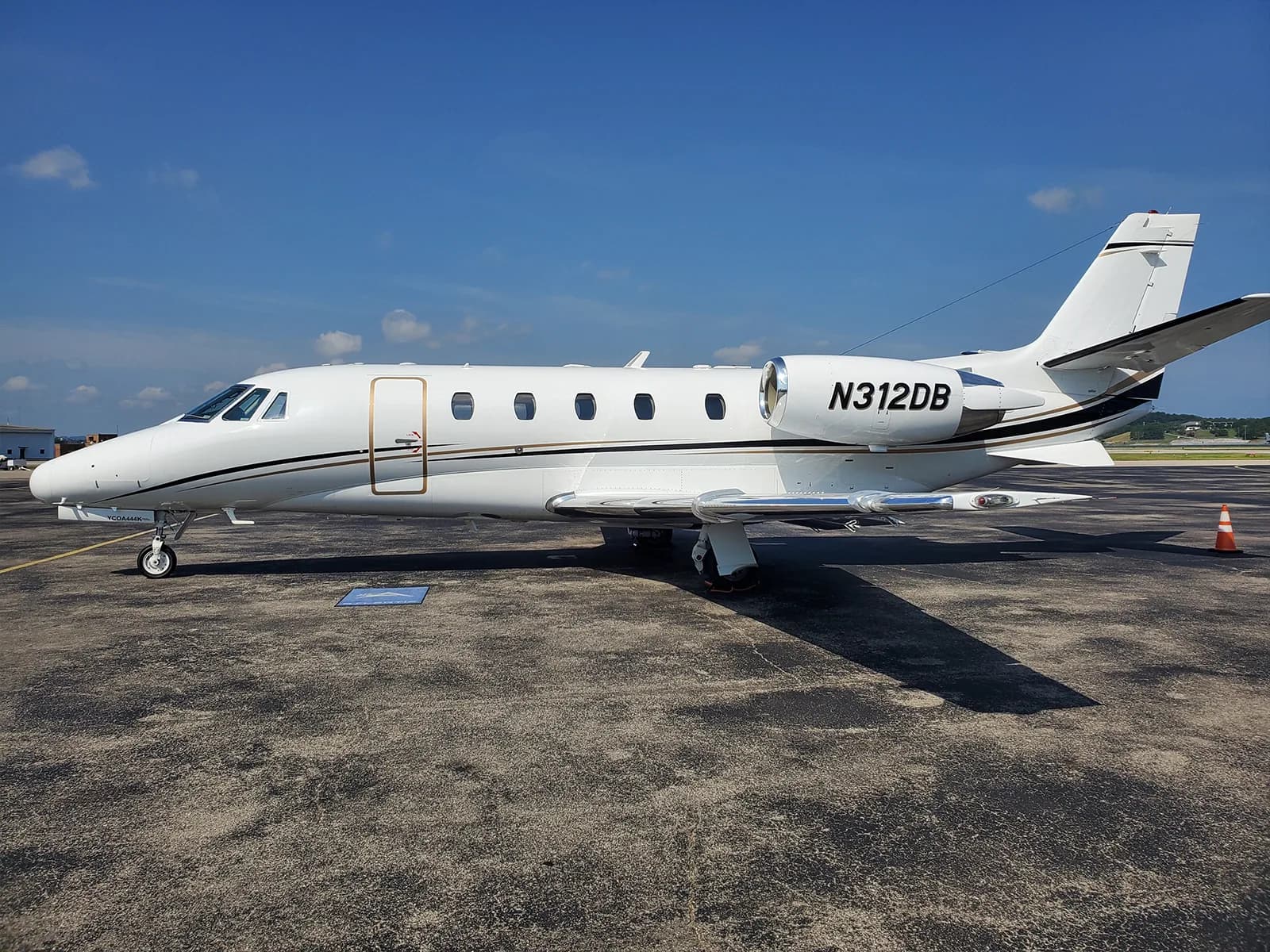 A white private jet with gold and black accent stripes, registration N312DB, parked on an asphalt tarmac under a blue sky.