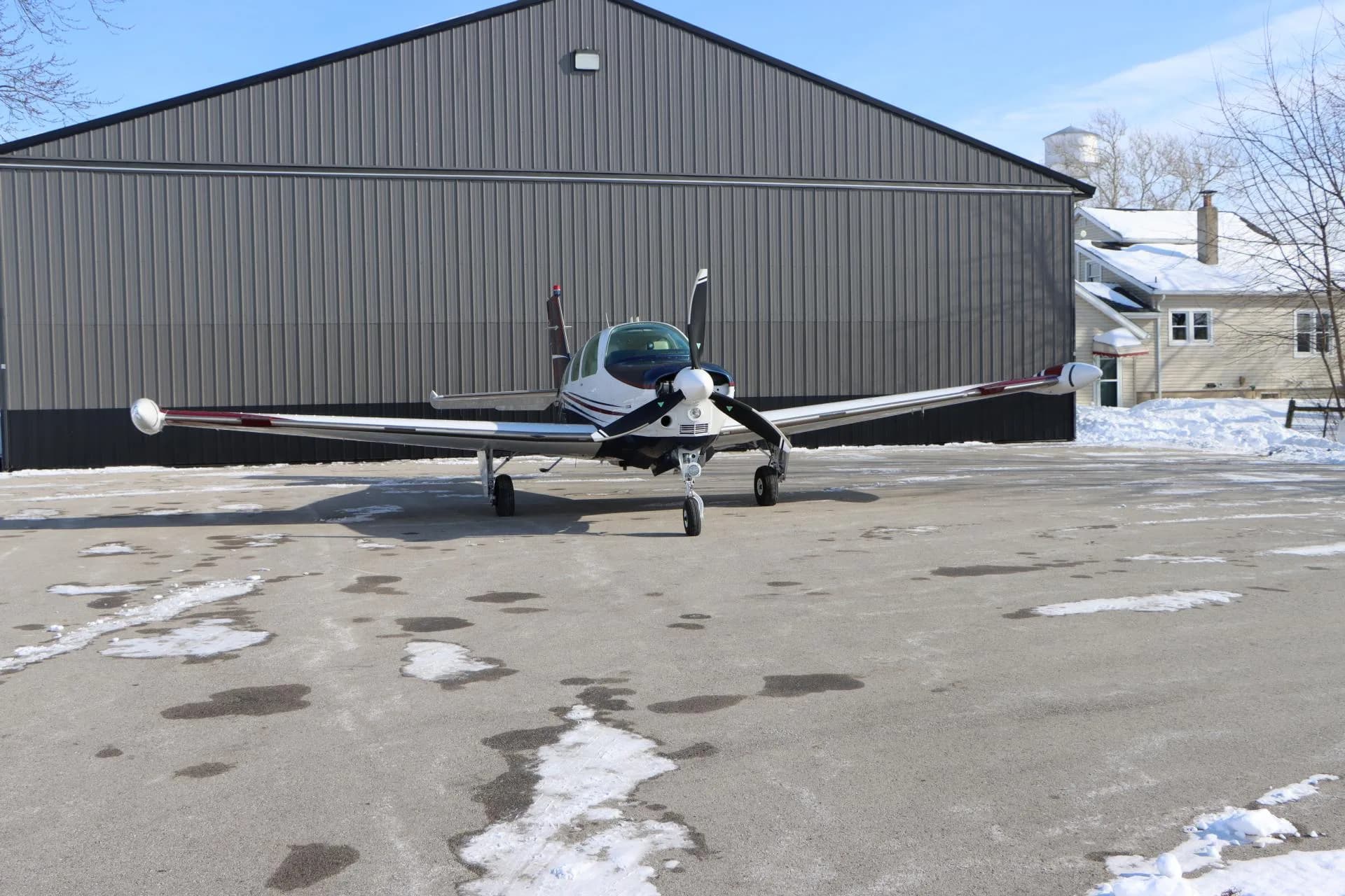 Front view of a white, blue, and red single-engine aircraft parked on a partially snowy tarmac in front of a hangar.