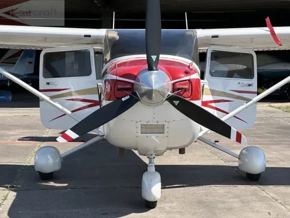 A white Cessna 182 Skylane with red and gold accents, seen from the front with its doors open on an airfield.