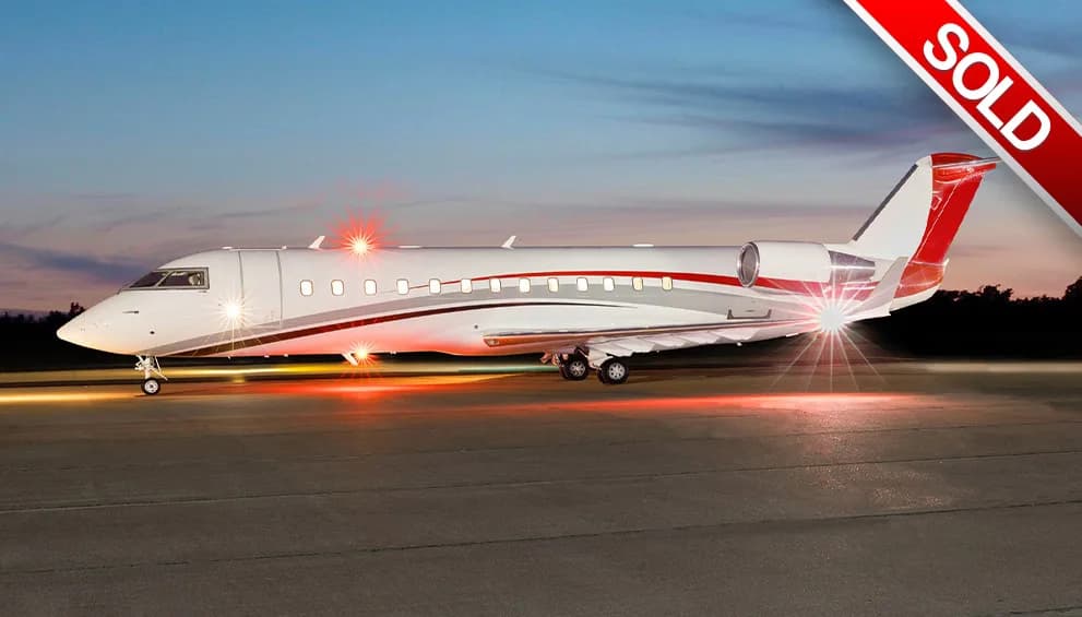 A white business jet with red and grey stripes sits on a tarmac at dusk with its lights illuminated.