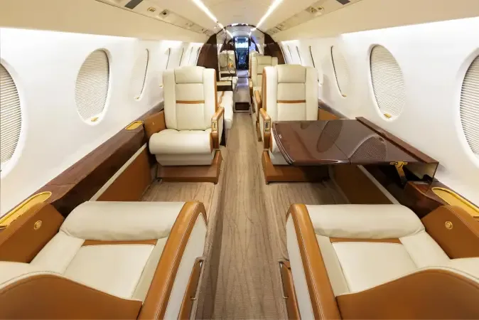 The luxurious interior cabin of a private jet, featuring cream leather seats, a dark wood table, and overhead lighting.