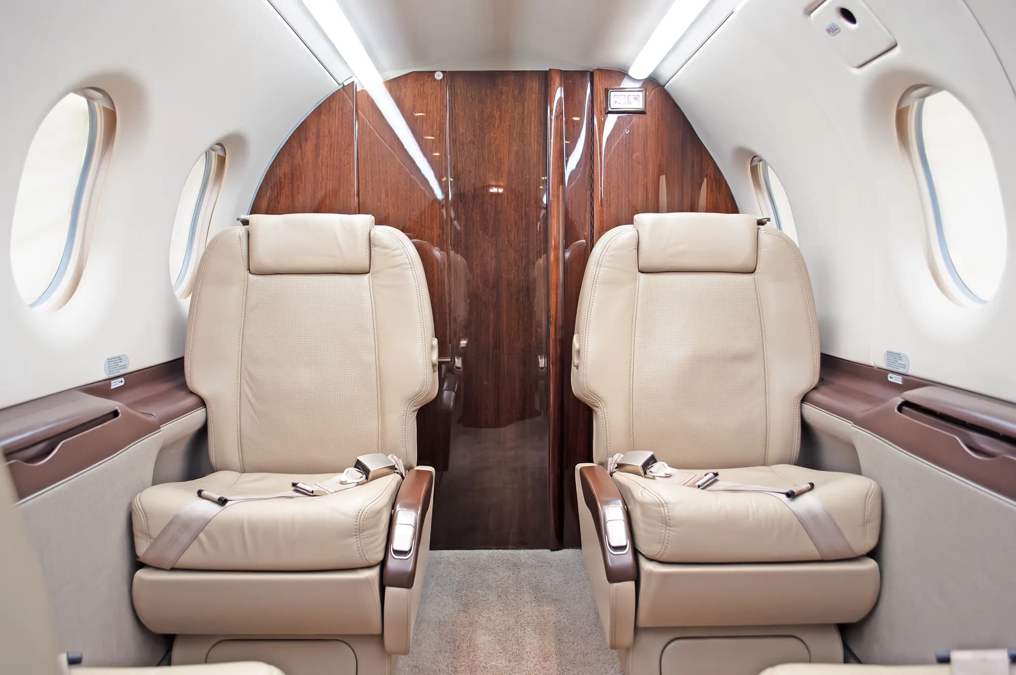 The interior cabin of a Pilatus PC-12 NG aircraft, showing two beige leather seats facing each other with a wooden bulkhead in the background.