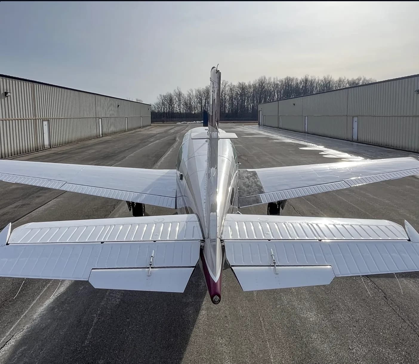 A rear view of a white and maroon Beechcraft Bonanza aircraft parked on a tarmac between two hangars.