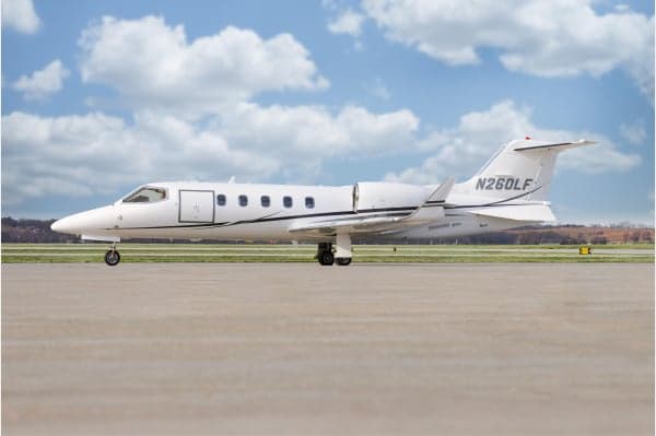 A white Learjet 75 business jet with black accent stripes is parked on an airfield under a partly cloudy sky.
