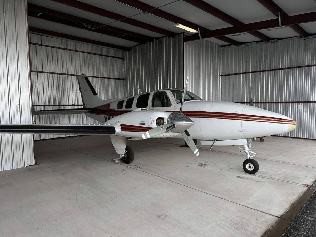A Beechcraft Baron aircraft with a white and maroon paint scheme is parked inside a hangar.