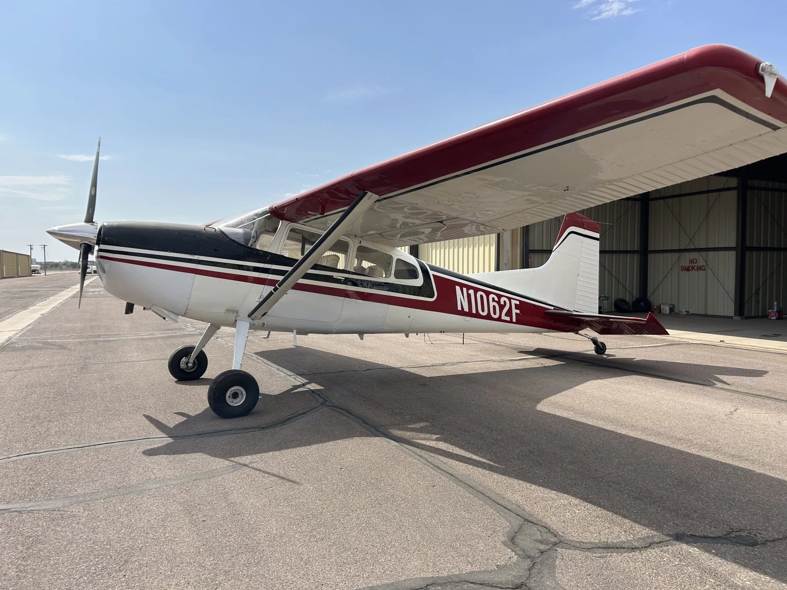 A white, black, and red Cessna 180 aircraft (N1062F) parked on an airport tarmac with a hangar in the background.