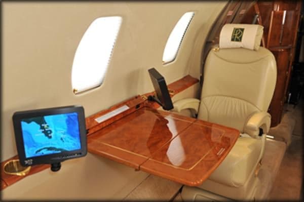 A cream leather private jet seat with a Rolls-Royce logo on the headrest is next to a polished wood table and an entertainment screen.