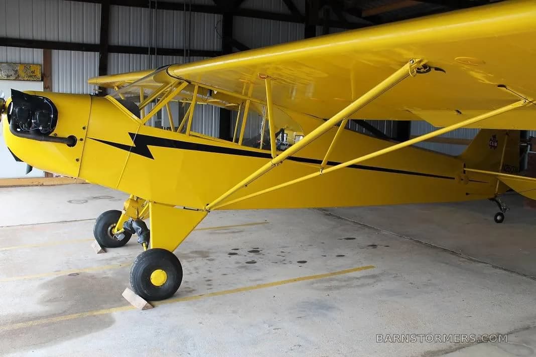 A yellow single-engine aircraft with black stripes, likely a Piper Cub, parked inside a hangar.