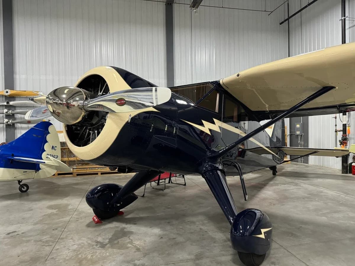 A dark blue and cream vintage aircraft with a lightning bolt design is parked in a hangar, showing its propeller and landing gear.