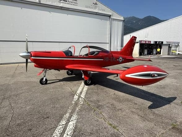 A red aircraft, HB-EML, parked on the tarmac in front of a hangar.