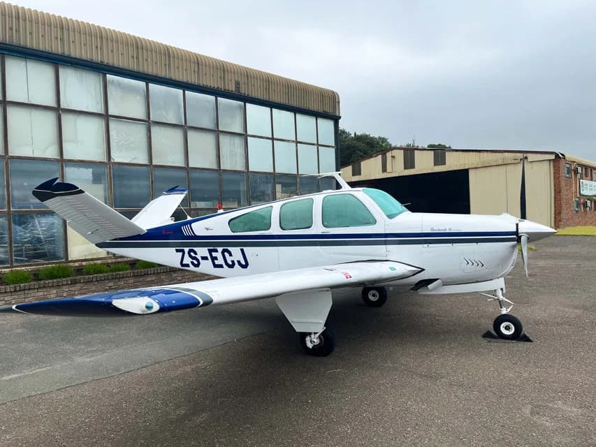 A white and blue Beechcraft Bonanza aircraft parked on the tarmac in front of a hangar.