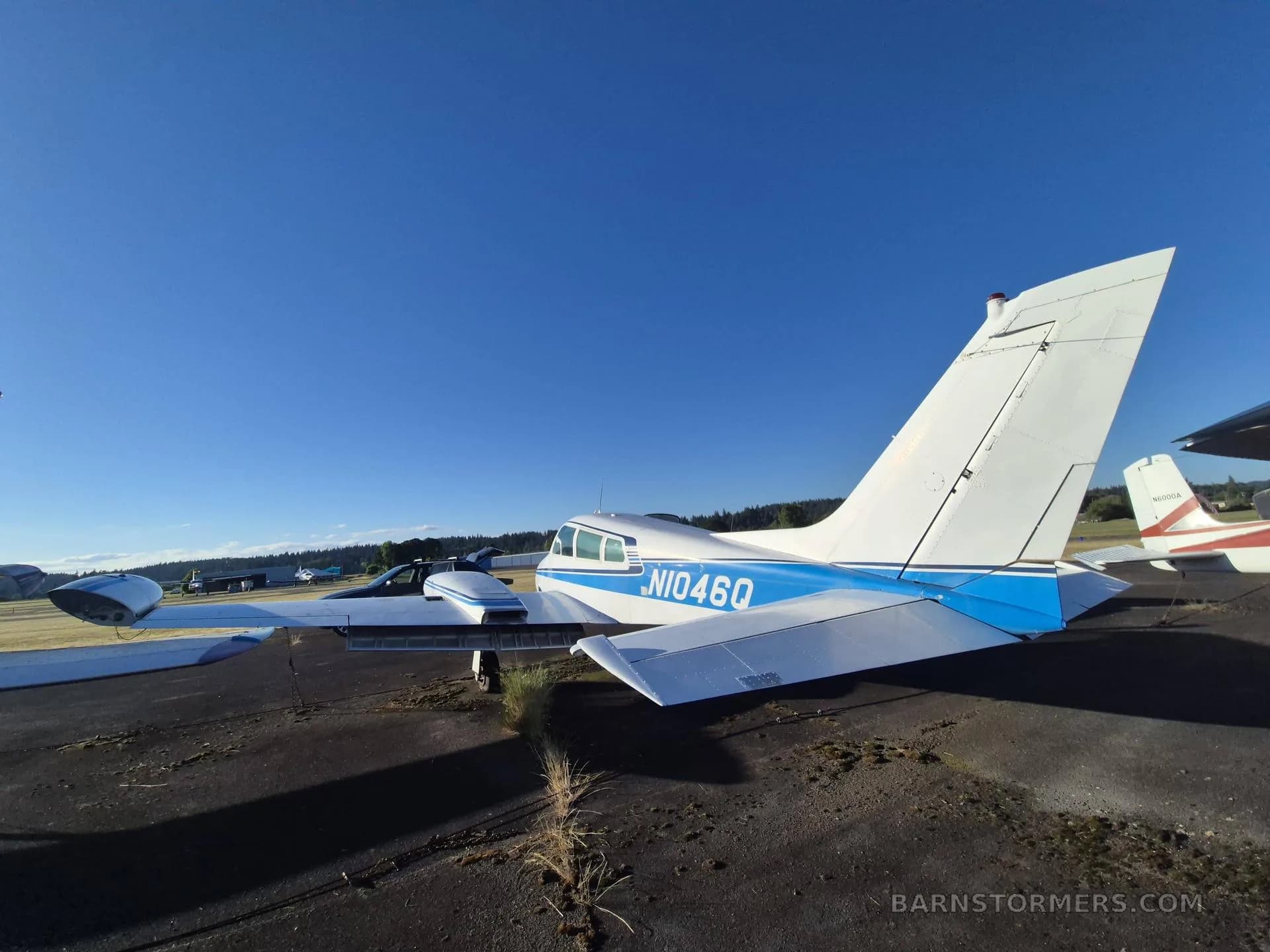 The rear-left view of a white twin-engine aircraft with blue stripes and registration N1046Q on its tail, parked on an asphalt ramp under a clear blue sky.