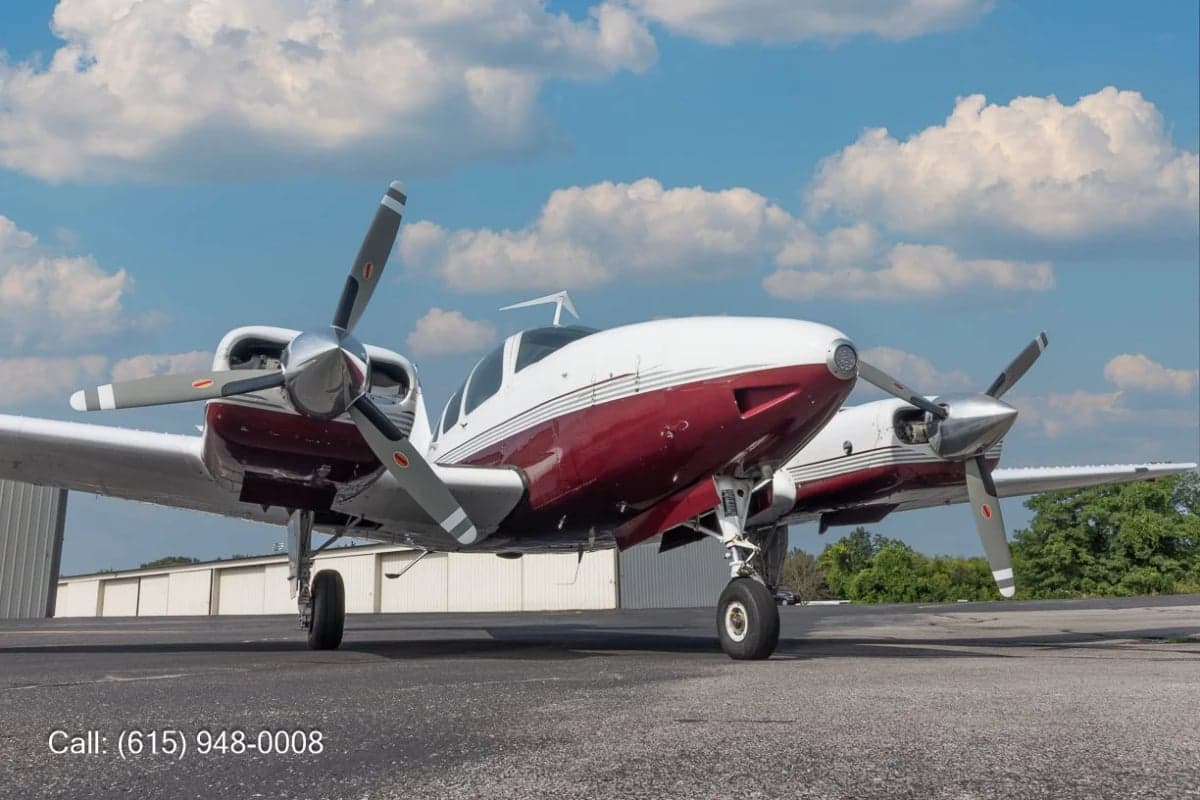 A front-quarter view of a white and red twin-engine propeller aircraft, likely a Beechcraft Baron, parked on an asphalt ramp under a blue sky.