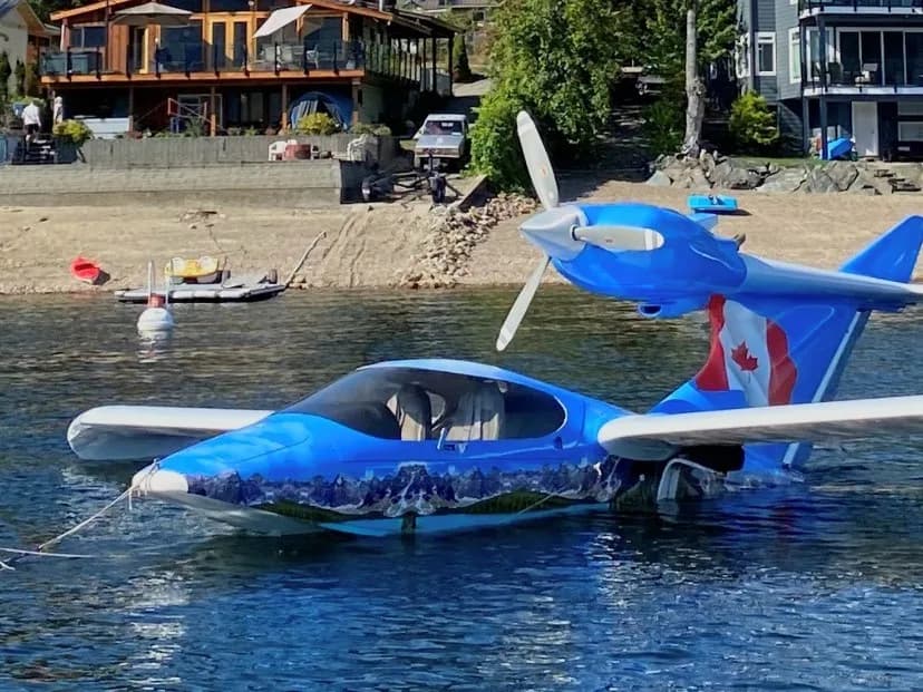 A blue amphibious aircraft with a mountain mural and Canadian flag on its tail floats in a lake, with houses on the shore in the background.