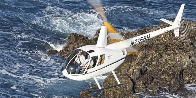 A white Robinson R44 helicopter flying over the ocean.