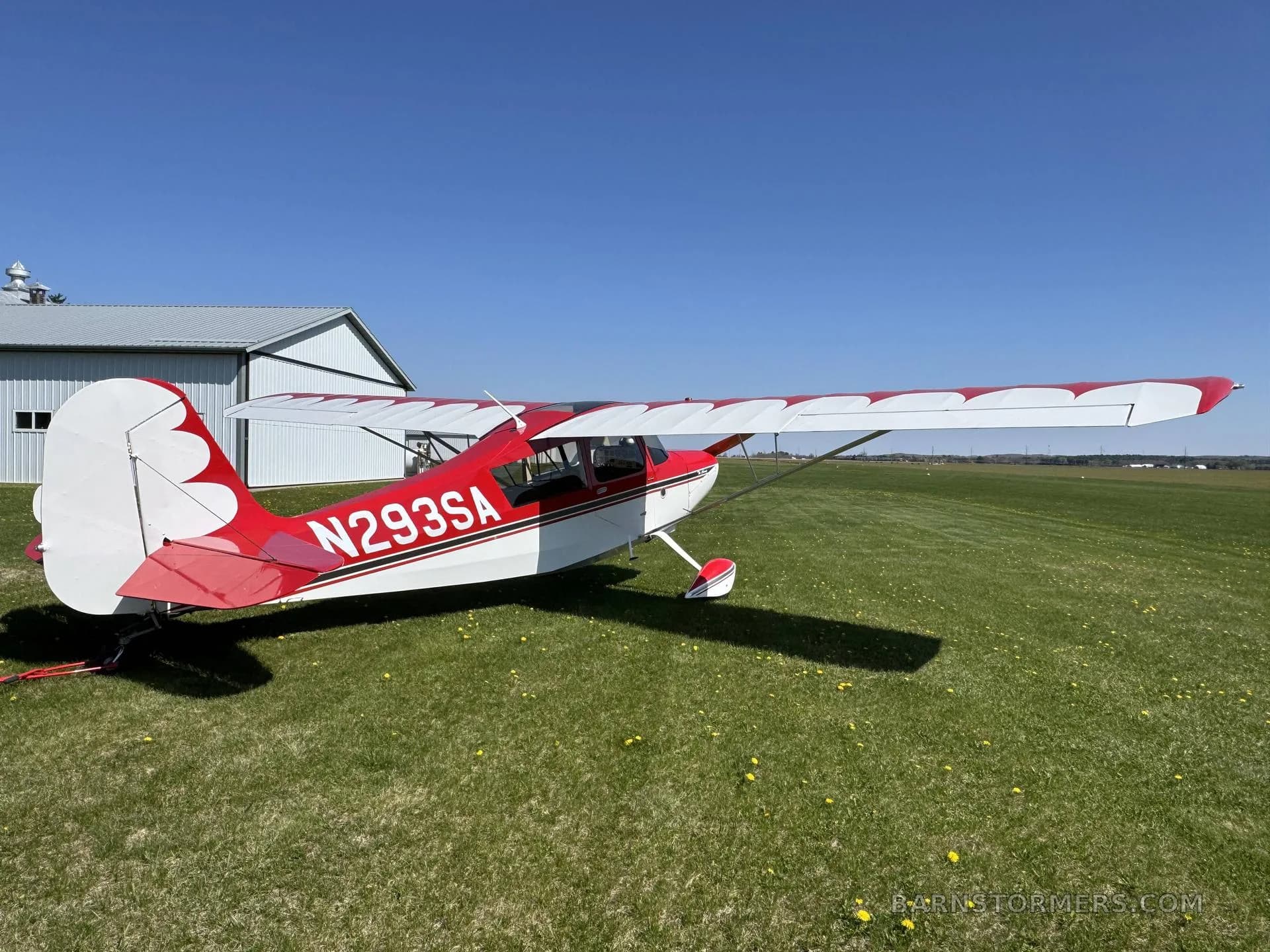 A red and white single-engine aircraft with tail number N293SA parked on a grassy field, showing its tail and a nearby hangar.