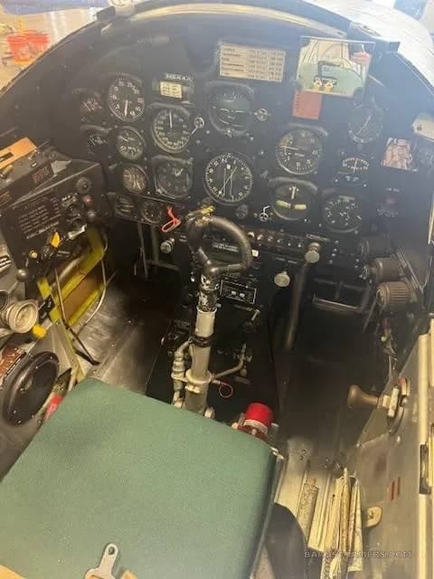 The detailed instrument panel and controls of a vintage aircraft cockpit, with a green seat cushion in the foreground.