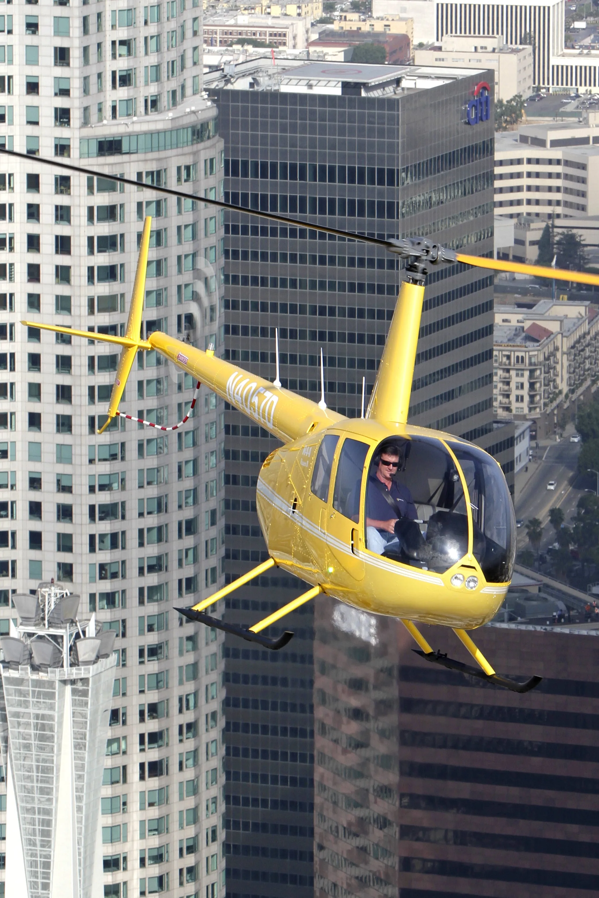 A yellow Robinson R44 helicopter flying in front of city buildings.