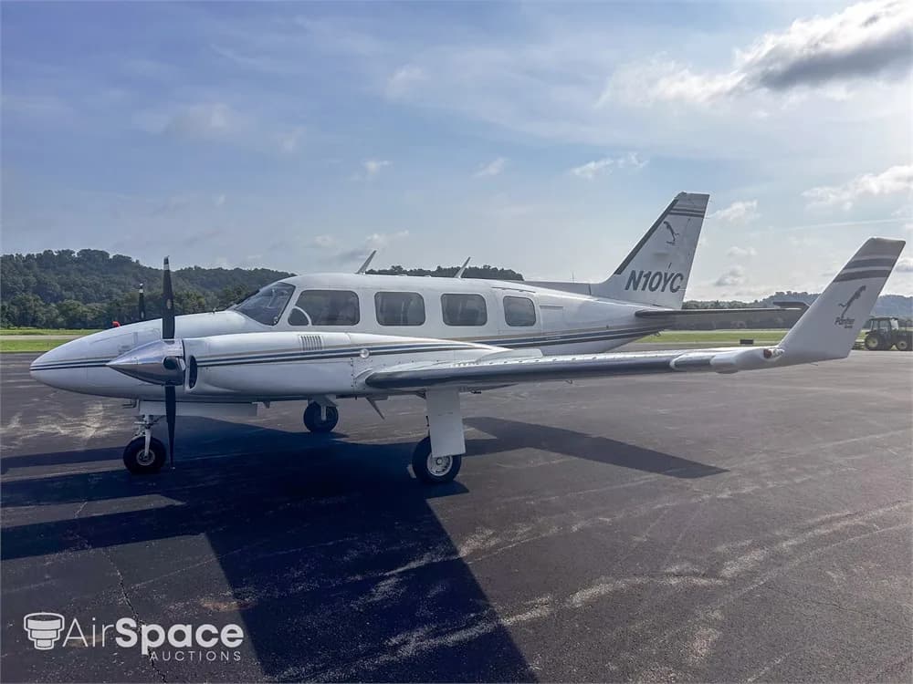 A white Piper Navajo aircraft with blue and gold stripes, registration N10YC, parked on an asphalt tarmac with hills in the background.