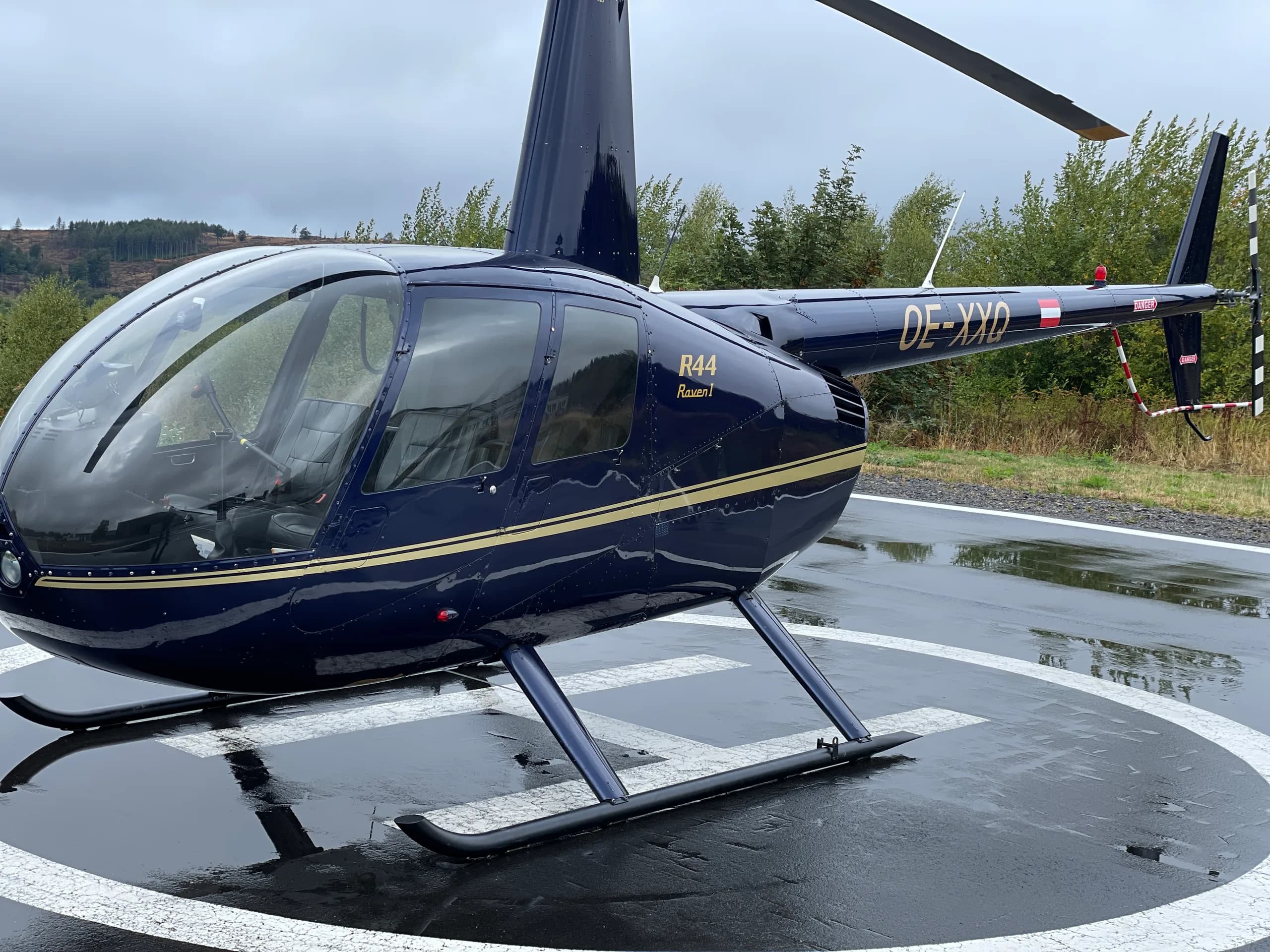 Close up of the Robinson R44 Raven I OE-XXQ helicopter on a helipad on a cloudy day.