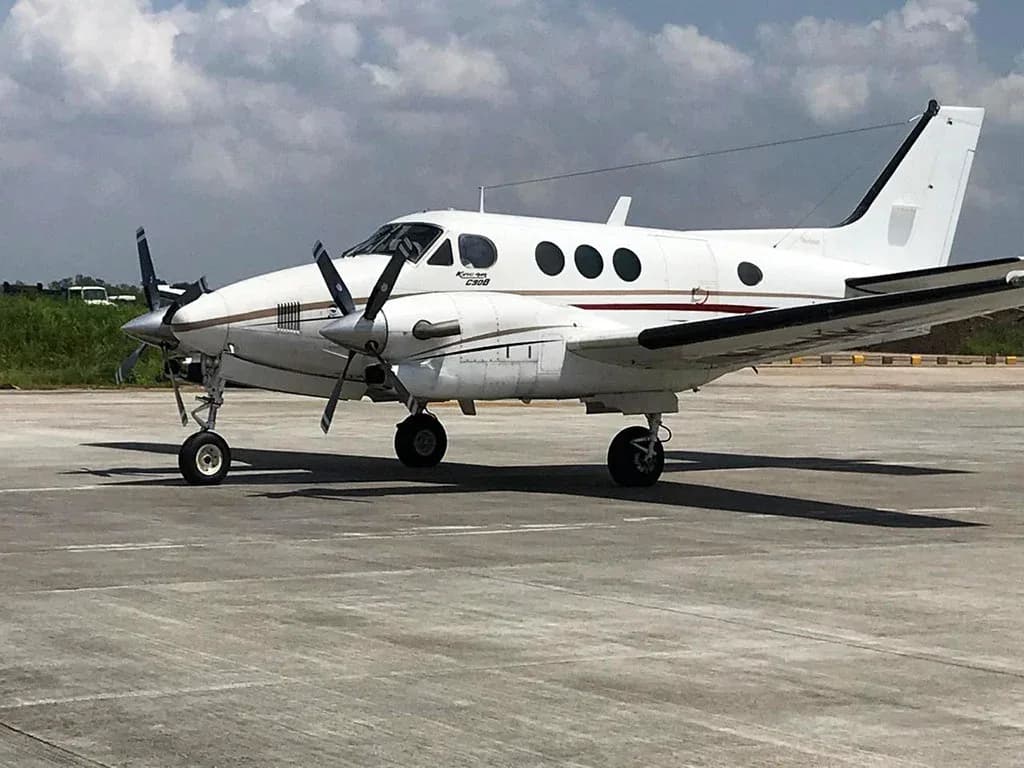 A white Beechcraft King Air C90B with brown and black stripes, parked on a concrete airfield under a partly cloudy sky.