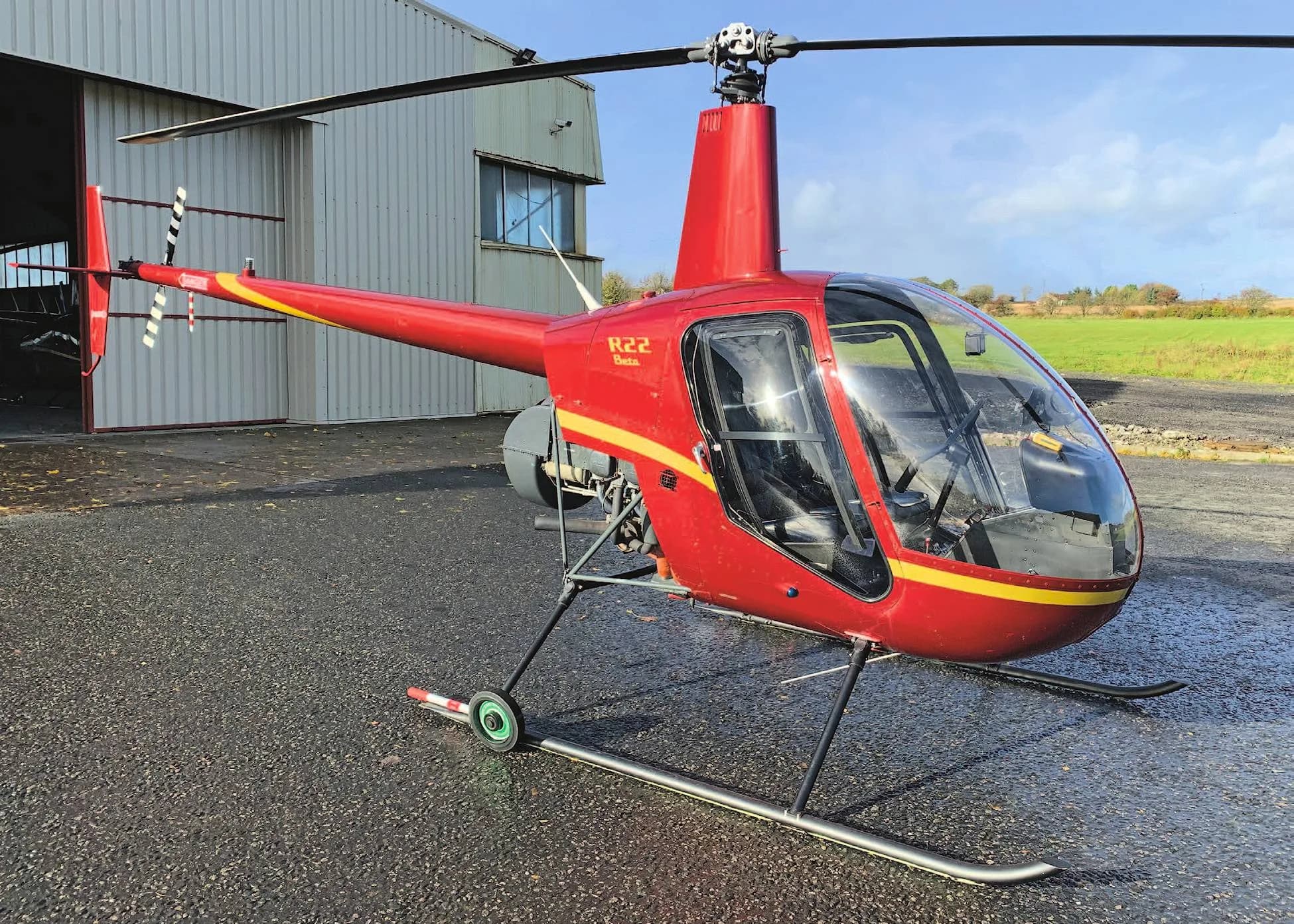 A red Robinson R22 Beta II helicopter with yellow stripes is parked on a wet tarmac in front of a hangar.