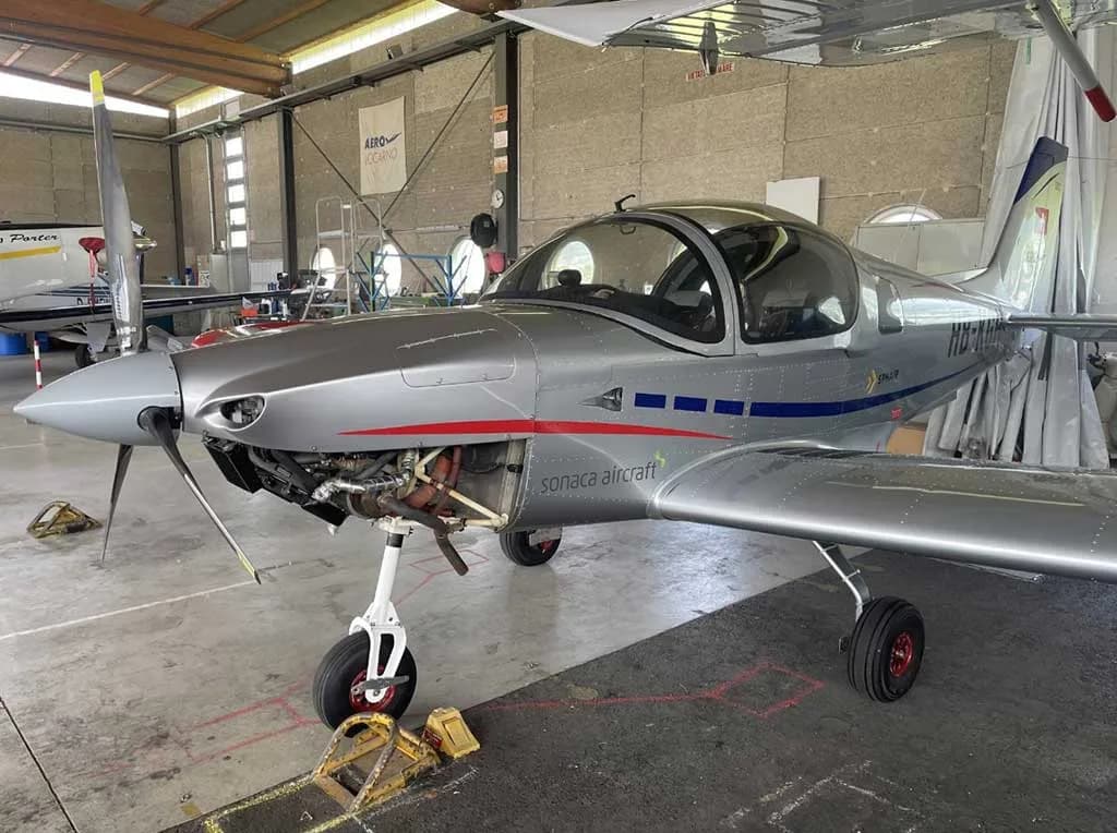 Three-quarter front view of a silver Sonaca 200 aircraft with blue stripes and open engine cowling, parked in a hangar.
