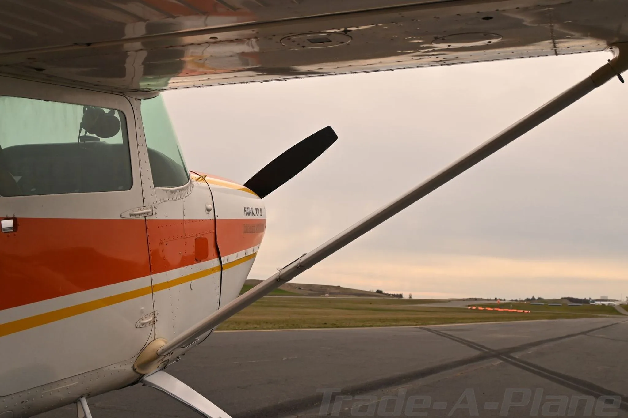 A close-up view of the front-left fuselage and wing of a white and orange Cessna 172 Hawk XP II (N5205V) on an airfield.