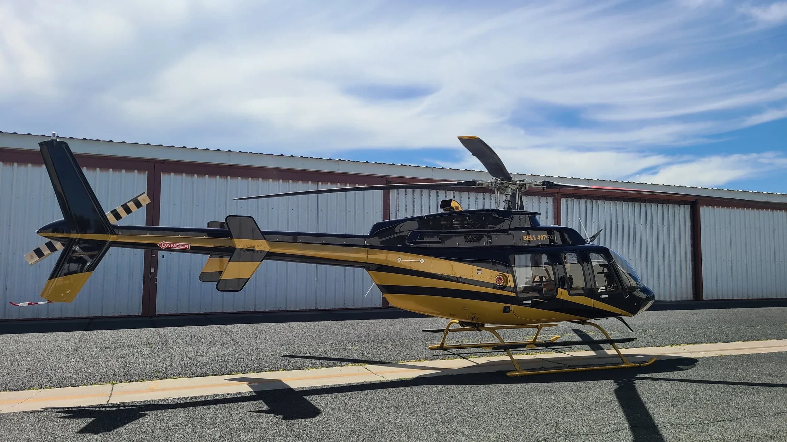 A black and gold Bell 407GX helicopter seen from the side, parked on an asphalt tarmac with a hangar in the background.