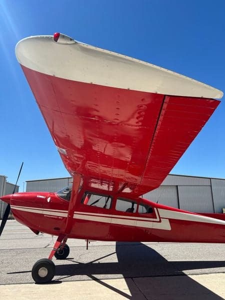 Side view of the red and white Cessna Skywagon 180 on the tarmac, with a hangar in the background.
