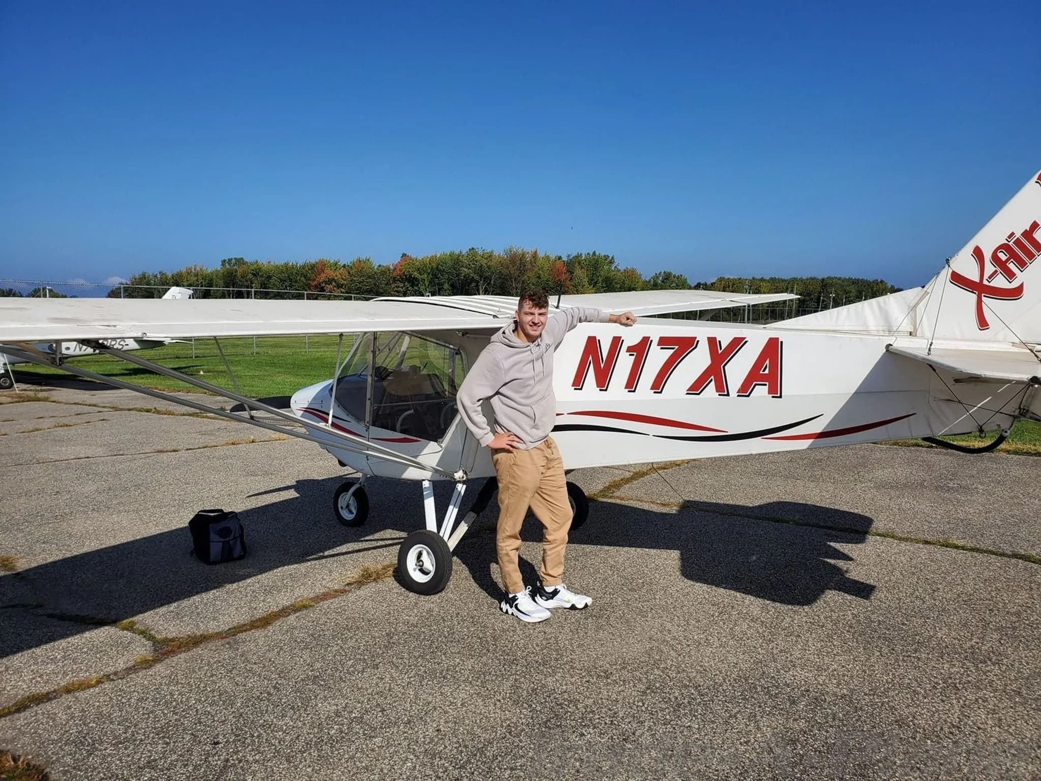 A man stands next to a white X-Air aircraft with red and black stripes, registration N17XA, on an airfield.