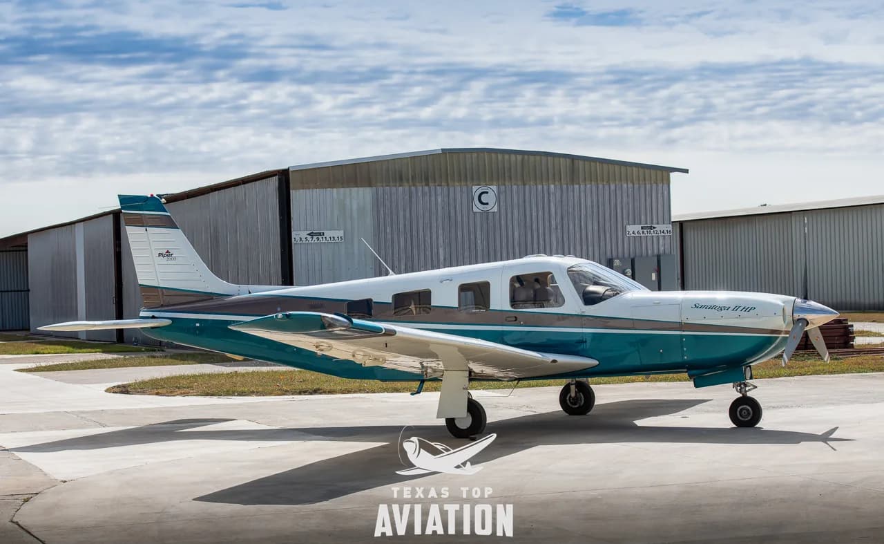 A white and teal Piper Saratoga II HP is parked in front of a hangar.