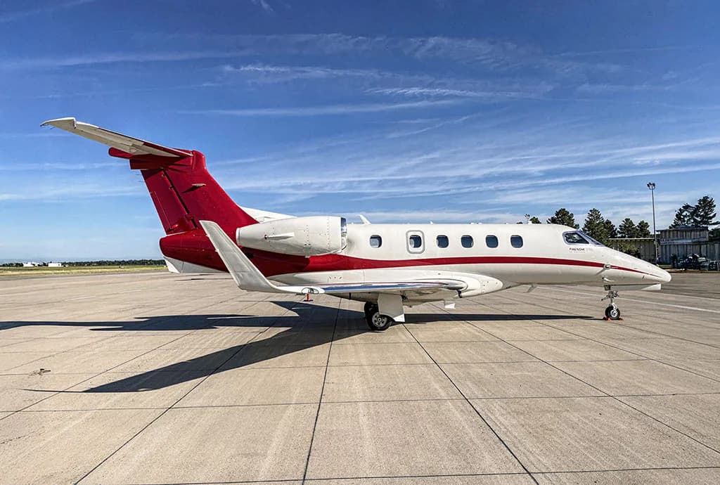 A white and red Embraer Phenom 300 private jet parked on an airport tarmac under a blue sky.