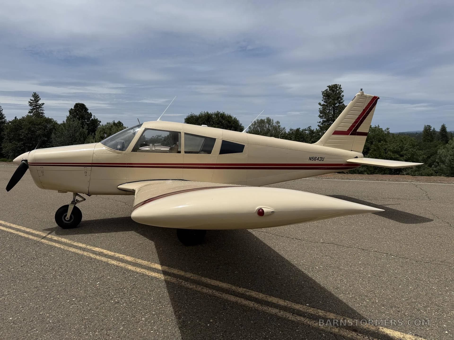 A cream-colored Piper Cherokee with red stripes, registration N5643U, parked on an asphalt surface under a partly cloudy sky.