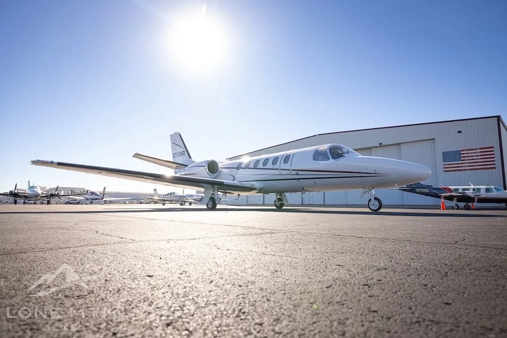 A low-angle side view of a white Cessna Citation Bravo jet, tail number N700NR, parked on an airport tarmac next to a hangar with an American flag.