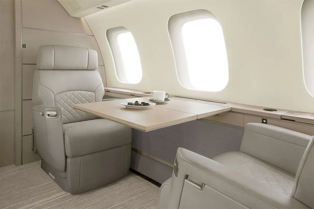 A private jet interior with two light grey seats facing a pull-out table set with a cup and pastries.