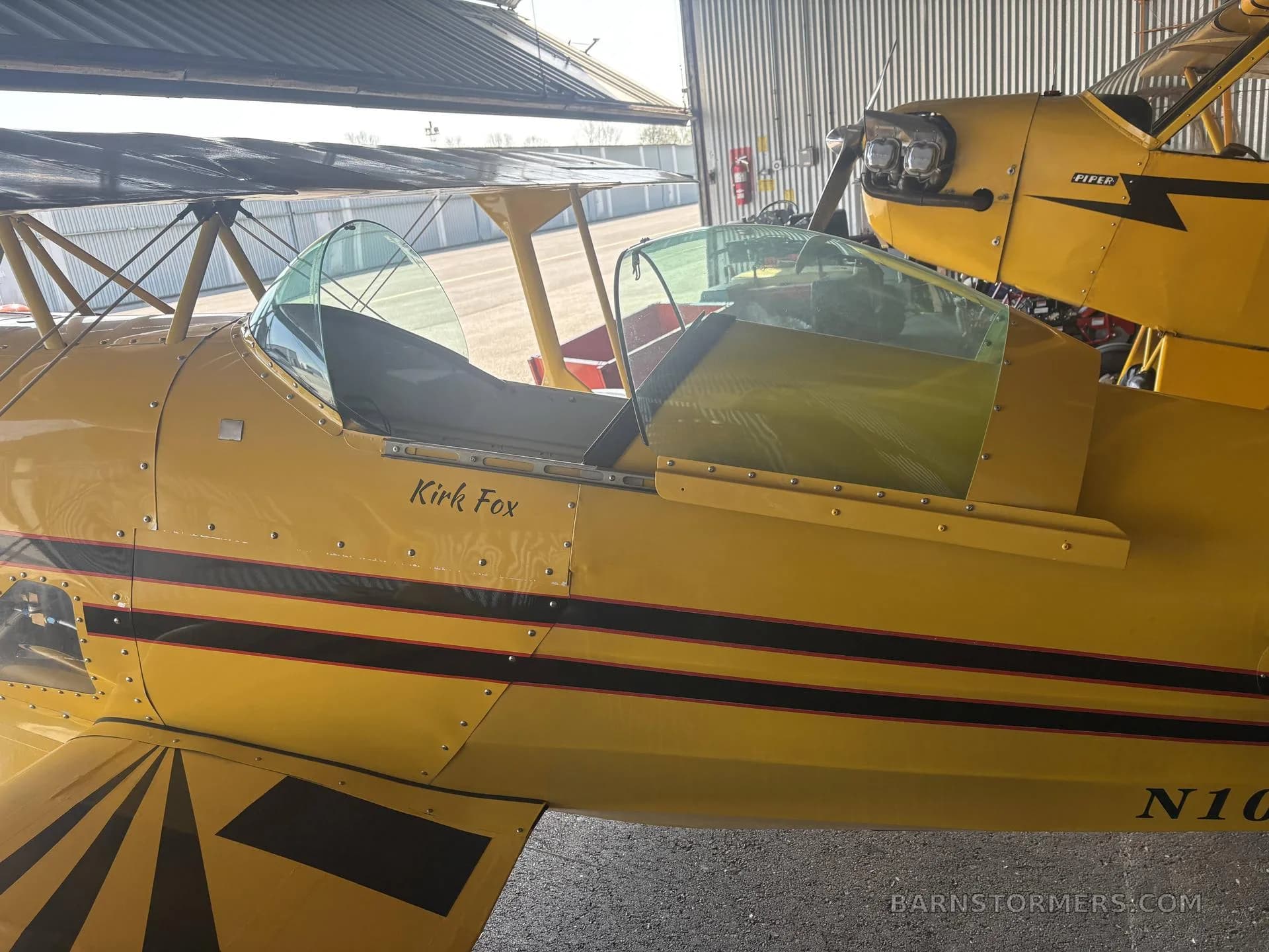 The side view of a yellow biplane with "Kirk Fox" painted on the fuselage, showing its open cockpits inside a hangar.