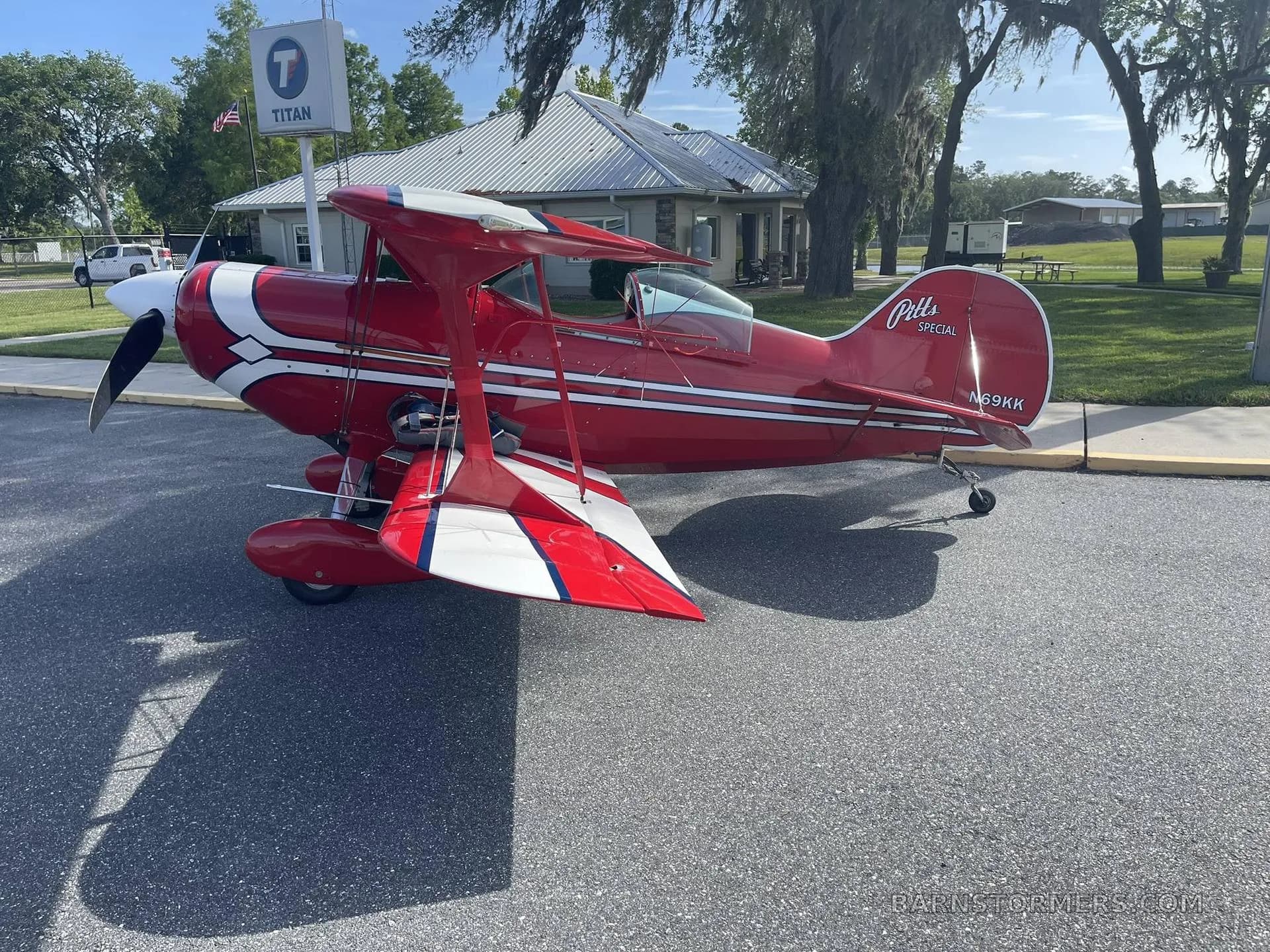 A red and white Pitts Special biplane, registration N69KK, is parked on an asphalt lot next to a building with a Titan sign.