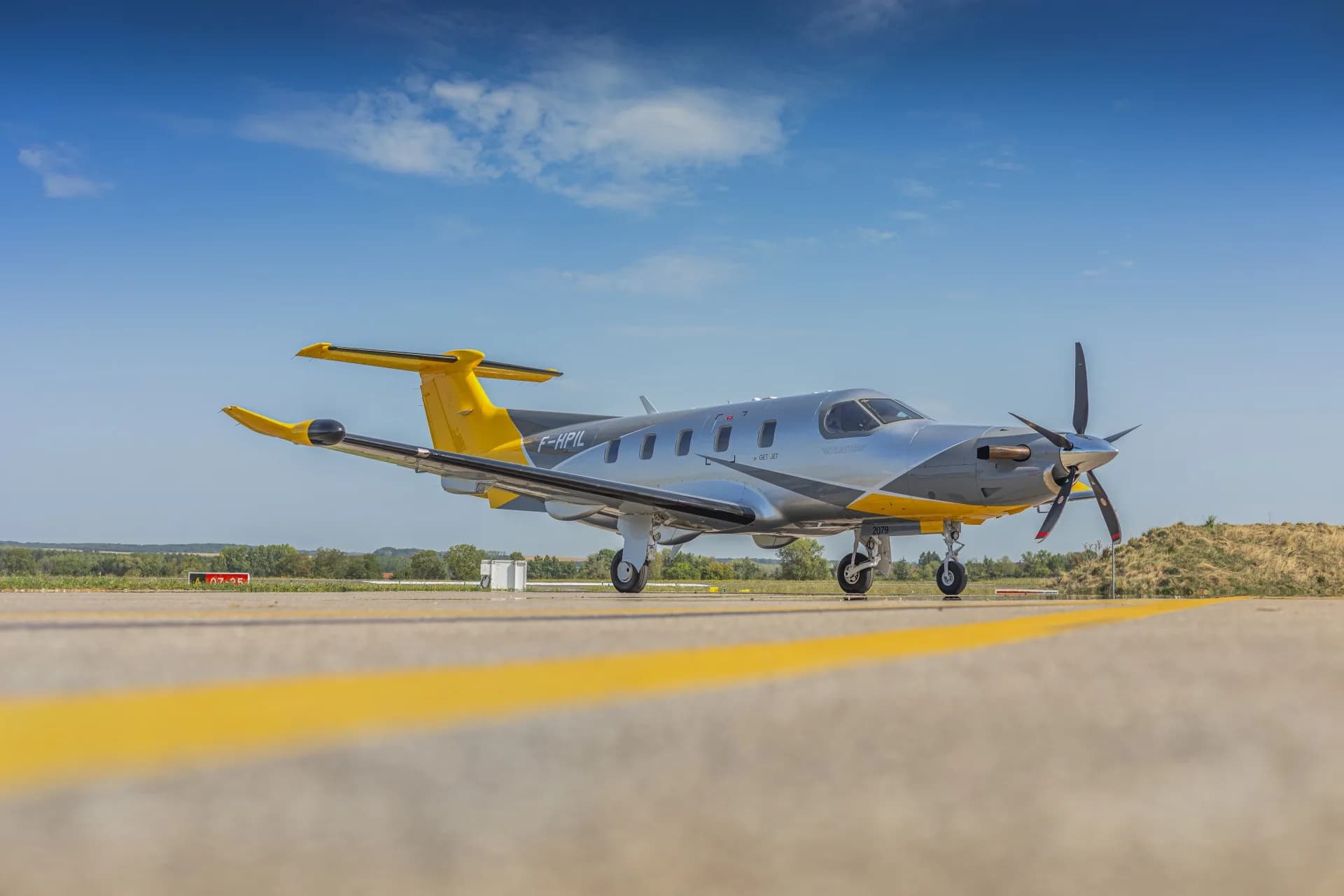 A low-angle view of the silver and yellow Pilatus PC-12 aircraft (F-HPIL) parked on an airfield with yellow markings.