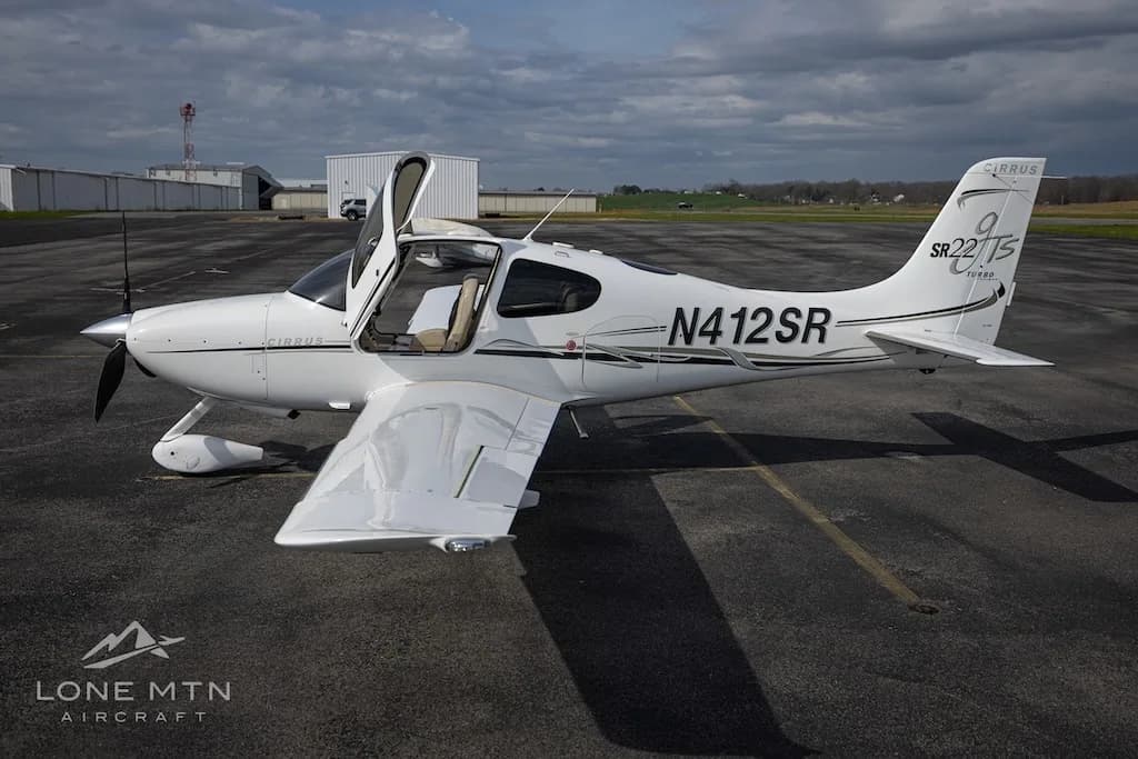 A white Cirrus SR22T aircraft with open doors is parked on an asphalt runway with hangars in the background.