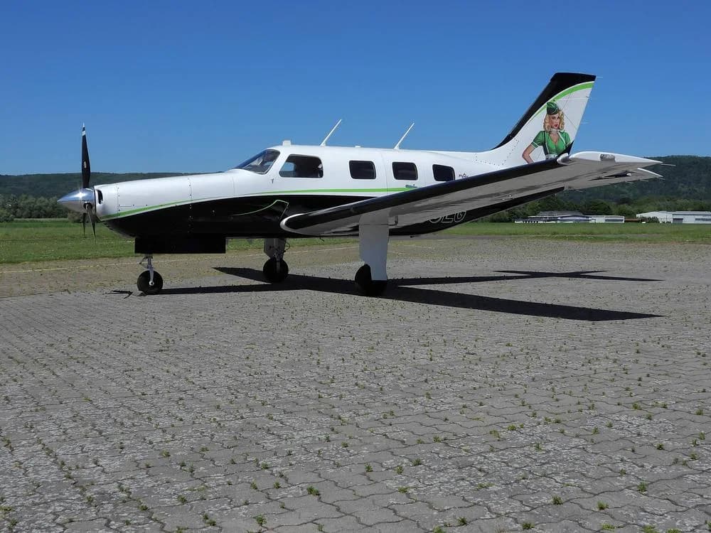 A side view of a white and black Piper M-Class aircraft with green accents and a pin-up girl on the tail, parked on a paved surface.