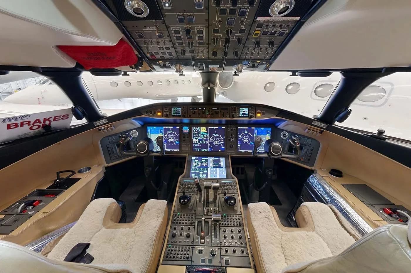 The modern cockpit of a business jet, featuring multiple large digital displays, a complex control panel, and two plush pilot seats.