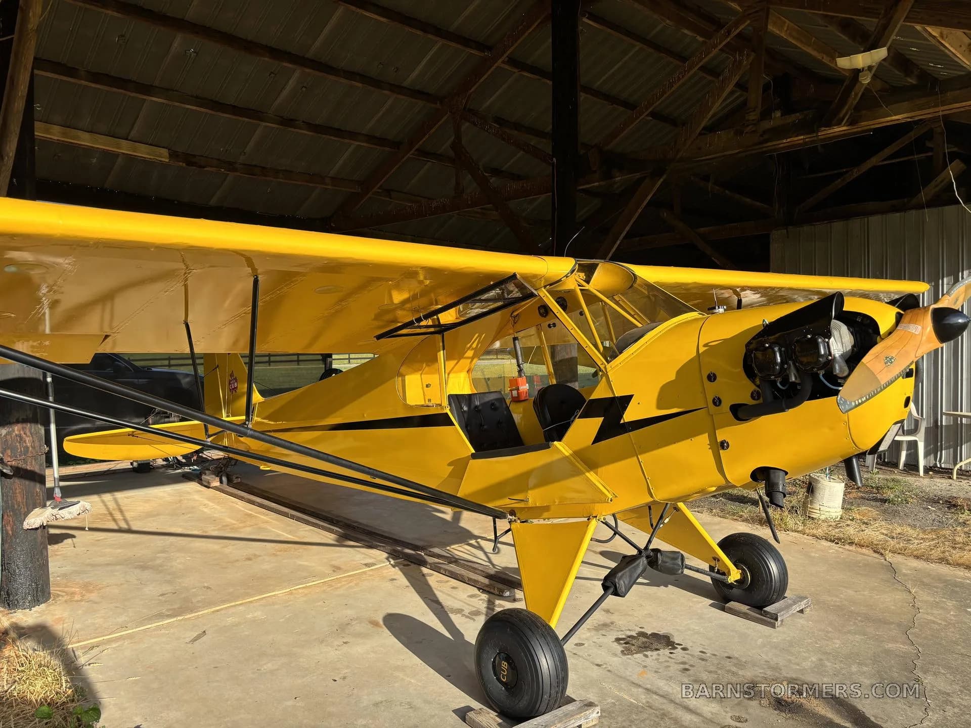 A yellow single-engine aircraft with an open cockpit door, parked on concrete inside a hangar with sunlight.