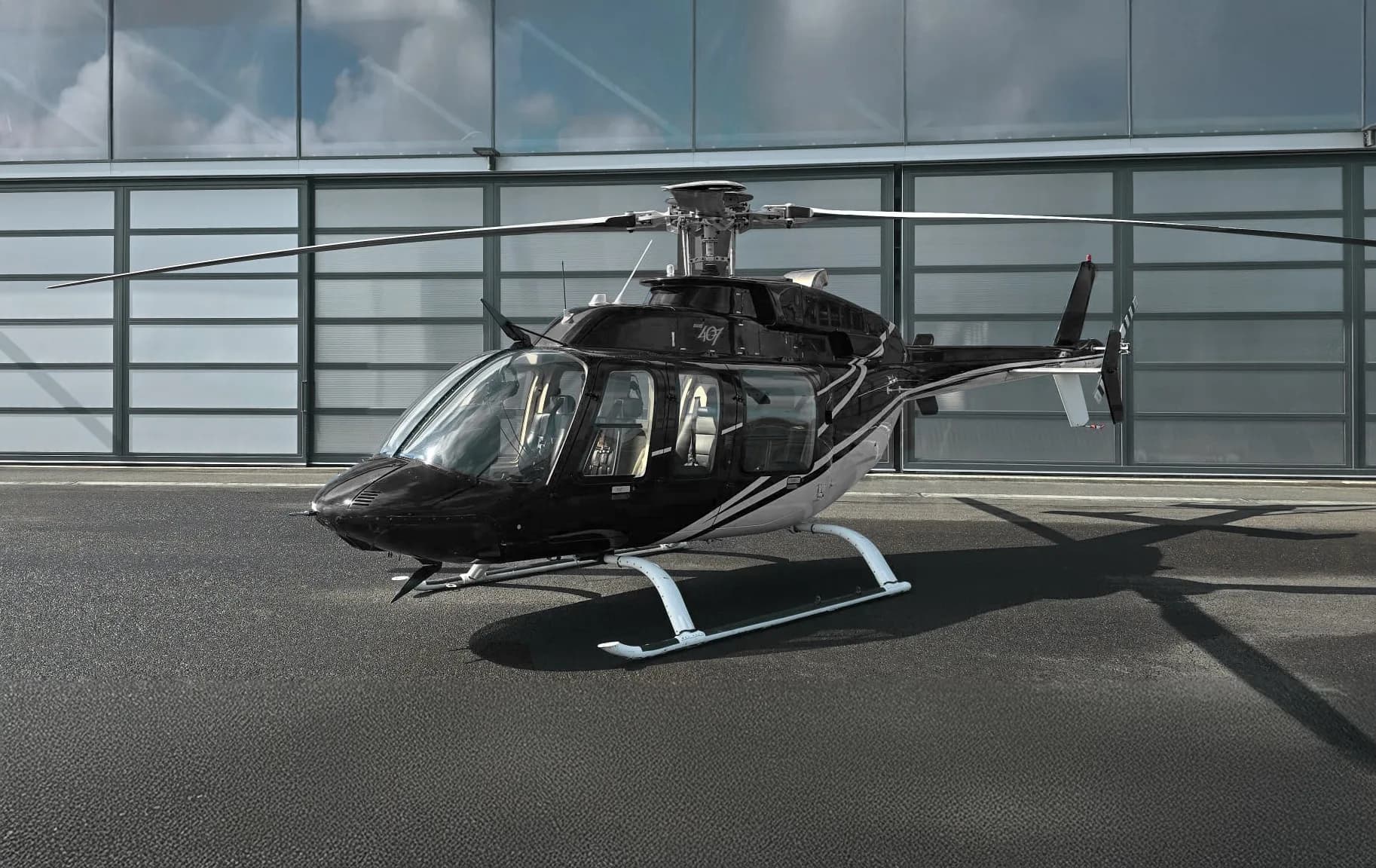 A black and silver helicopter is parked on an asphalt apron in front of a modern hangar.
