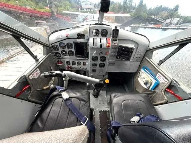 The cockpit of a floatplane with a detailed instrument panel, two pilot seats, and a view of the water outside.