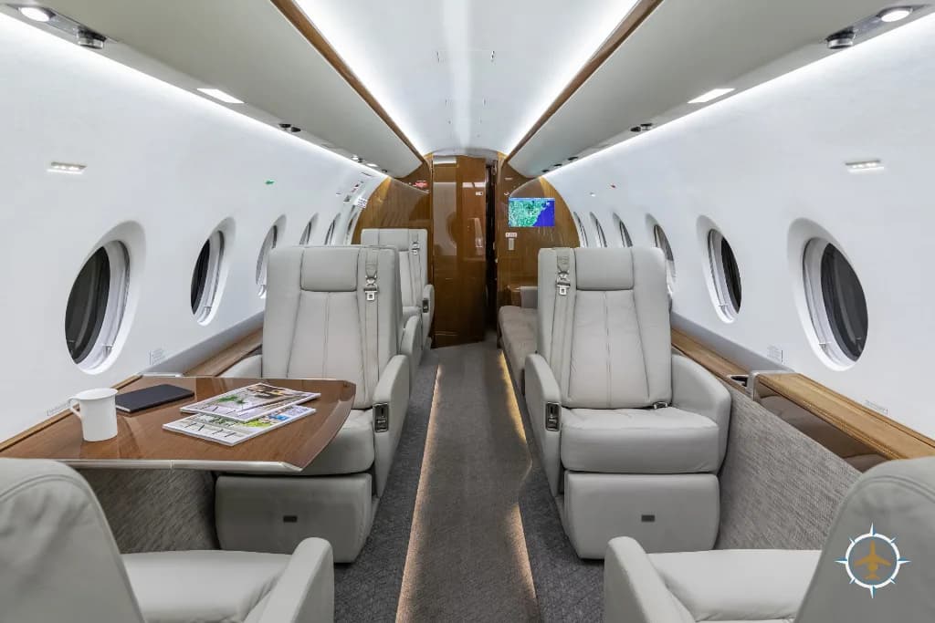 The luxurious aircraft cabin with light grey leather seats, a wooden table, and large windows.